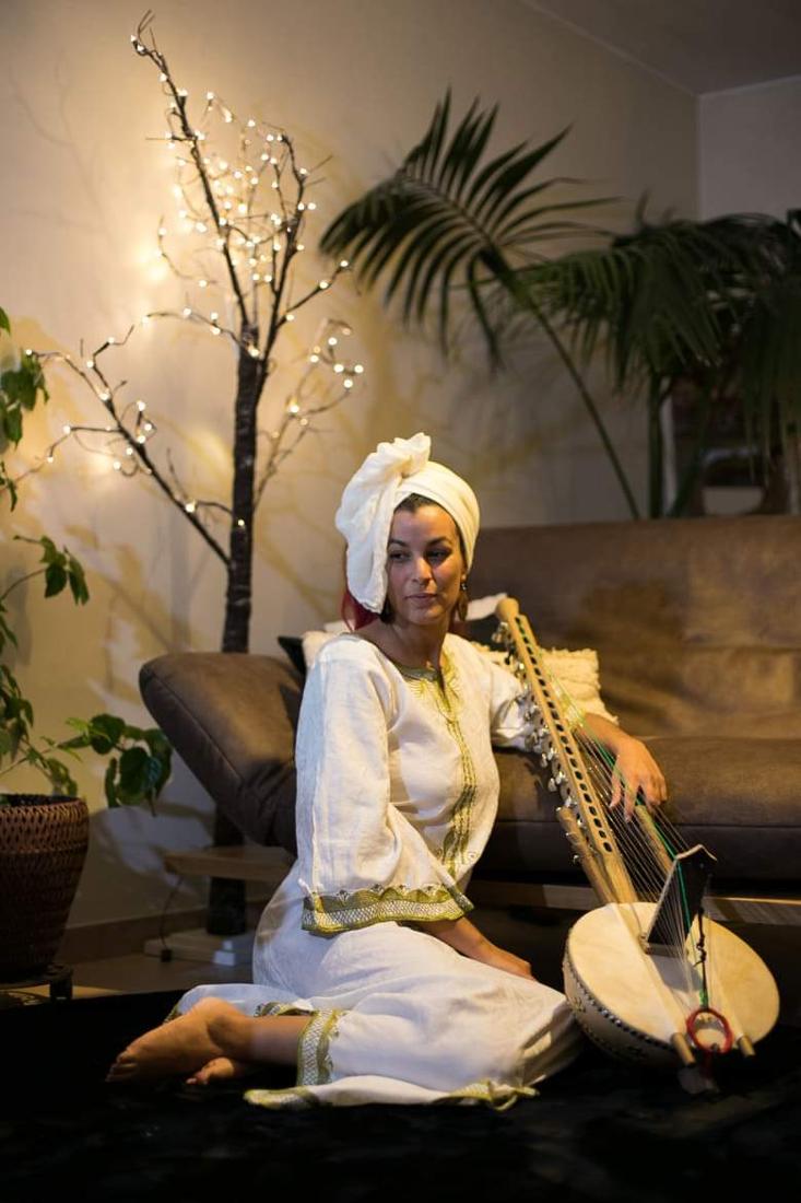 Maïya in white, seated beside her kora, among plants and soft lights