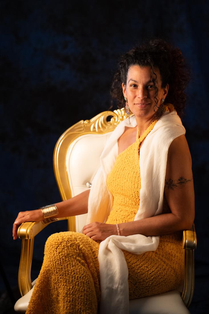 Maïya Katy seated on a gilded chair, in a saffron dress and ivory shawl